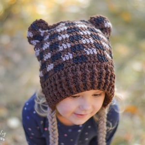 May include: A child wearing a brown and tan crocheted bear hat with ear flaps. The hat has a checkered pattern and is pulled down over the child's forehead.