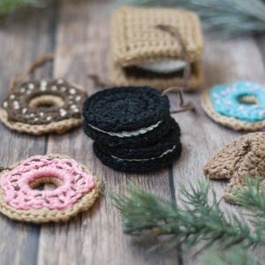 May include: Crocheted food ornaments, including a donut with pink icing, a donut with brown icing, a stack of chocolate chip cookies, a blue iced donut, and a fortune cookie.