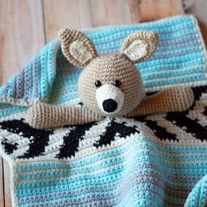 May include: A crocheted baby blanket with a brown and white crocheted animal toy in the centre. The blanket has a blue, grey, and white striped pattern and a black and white geometric pattern.