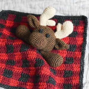 May include: A brown crocheted moose toy laying on a red and black plaid crocheted blanket.