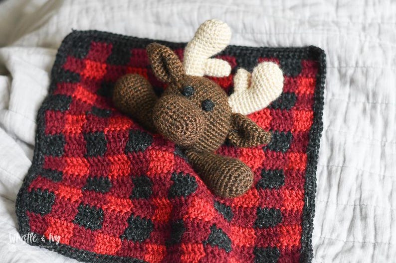 May include: A brown crocheted moose toy laying on a red and black plaid crocheted blanket.