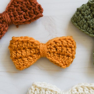 Puede incluir: Primer plano de varios lazos de ganchillo hechos a mano en varios colores: naranja, óxido, verde oliva y crema. Los lazos están dispuestos sobre una superficie blanca. Se aprecia la textura del hilo.