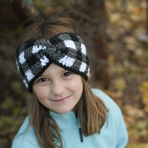 May include: A black and white checkered crochet headband with a twisted design. The headband is worn by a young person with brown hair.