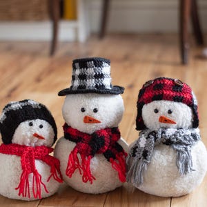 May include: Three handmade snowmen with white bodies, each wearing a different crocheted hat and scarf. The hats are black and white, red and black plaid, and black and red plaid. The scarves are red and black, and gray and black.