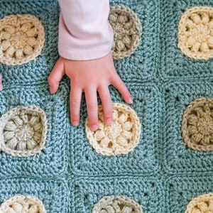 May include: A baby's hand rests on a crocheted baby blanket with a blue background and multi-colored squares. The blanket features a repeating pattern of crocheted flowers.