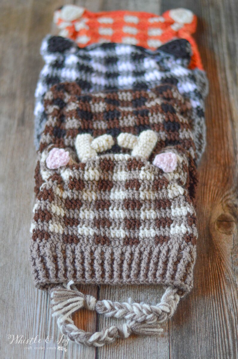 May include: Three crocheted hats stacked on top of each other. The top hat is orange with white polka dots, the middle hat is black and white checked, and the bottom hat is brown and white with a braided tassel.
