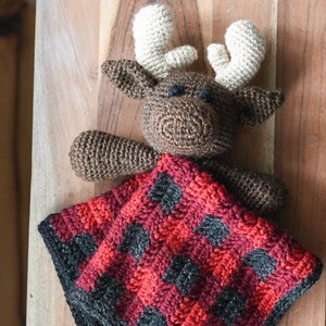 May include: A brown crocheted moose with white antlers holding a red and black plaid crocheted baby blanket. The blanket has a buffalo plaid pattern.