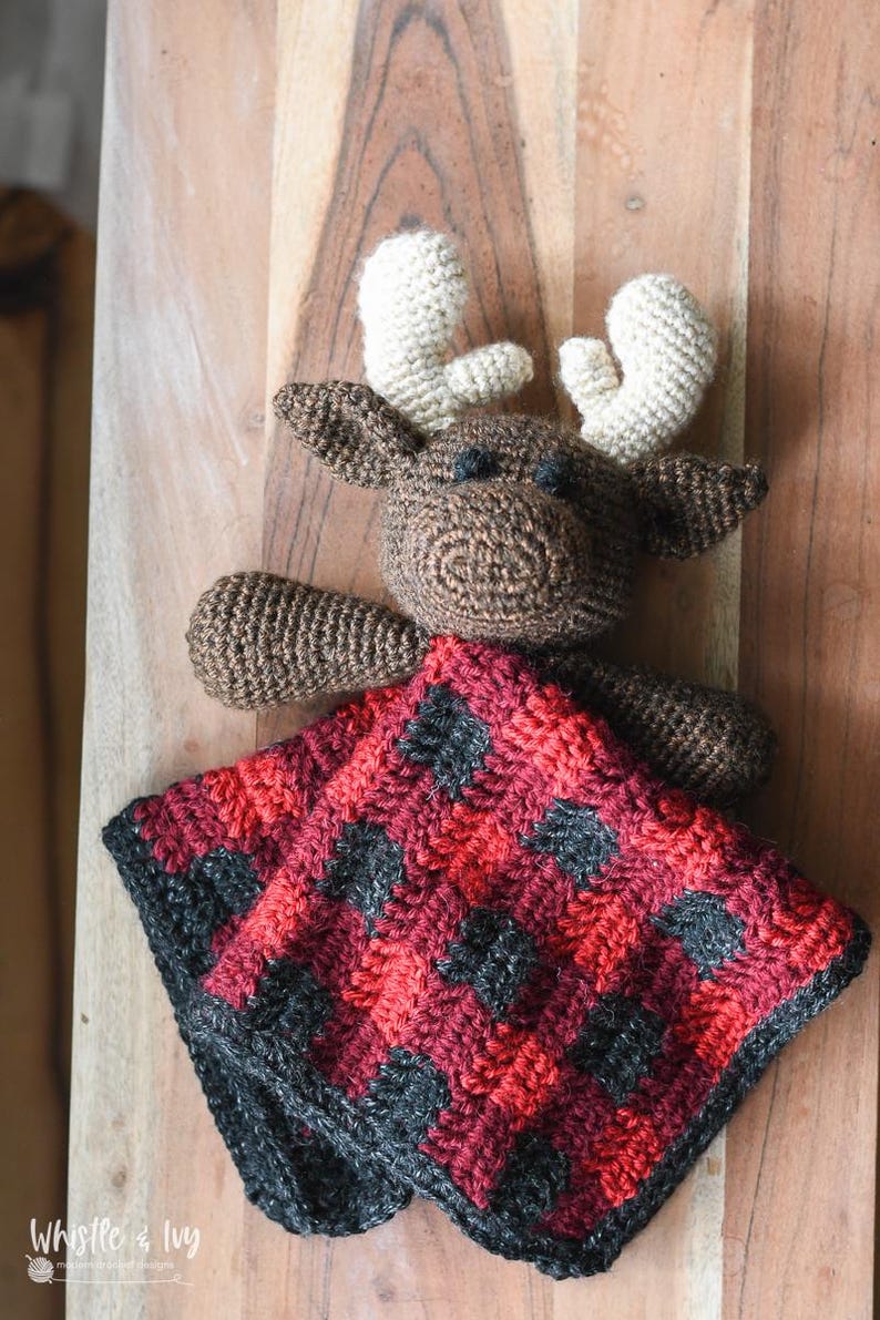 May include: A brown crocheted moose with white antlers holding a red and black plaid crocheted baby blanket. The blanket has a buffalo plaid pattern.