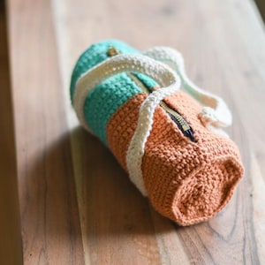 May include: A handmade crochet bag in shades of teal, orange, and cream. The cylindrical bag features a zip closure and a cream-coloured strap. The bag is made of textured yarn and is lying on a wooden surface.