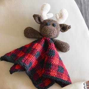 May include: A crocheted moose stuffed animal with a red and black plaid blanket. The moose has brown fur and white antlers.