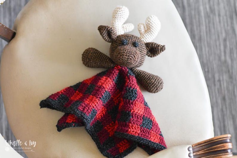 May include: A crocheted moose stuffed animal with a red and black plaid blanket. The moose has brown fur and white antlers.