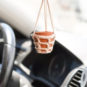 May include: A small terracotta pot with a crocheted cover hangs from a string in the interior of a car. The pot is hanging from the rearview mirror.