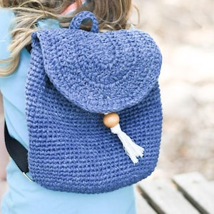May include: A small blue crocheted backpack with a wooden bead and tassel. The backpack is worn by a child.