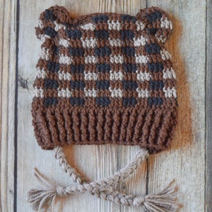May include: A brown, black and beige crocheted hat with bear ears and a braided cord. The hat is on a wooden background.