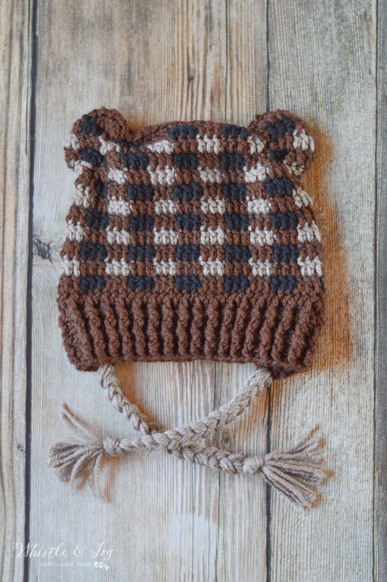 May include: A brown, black and beige crocheted hat with bear ears and a braided cord. The hat is on a wooden background.