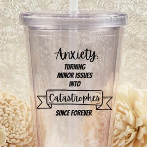 May include: A clear plastic tumbler with a white straw and lid. The tumbler has the words "Anxiety: Turning Minor Issues Into Catastrophes Since Forever" printed in black. The background features cream-colored flowers.