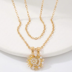 May include: Gold-toned necklace set featuring a sun and moon pendant. The pendants are adorned with clear, sparkling crystals. The necklace has two chains, one shorter and one longer, both with a delicate link design. The jewelry is displayed on a white surface.