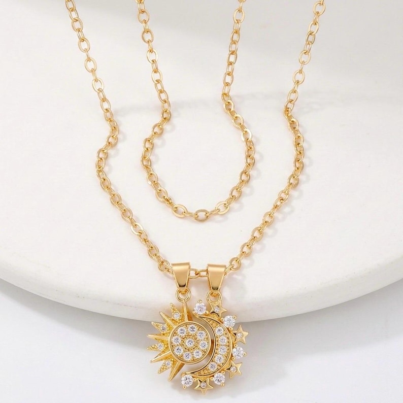 May include: Gold-toned necklace set featuring a sun and moon pendant. The pendants are adorned with clear, sparkling crystals. The necklace has two chains, one shorter and one longer, both with a delicate link design. The jewelry is displayed on a white surface.