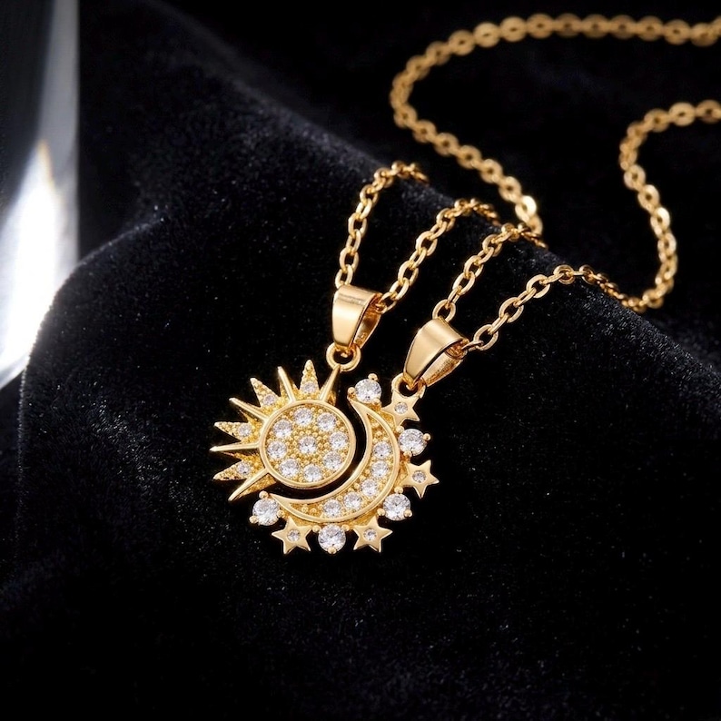 May include: Gold-toned necklace featuring two pendants: a sun and a crescent moon. The sun pendant has a circular center with small, clear stones and radiating rays. The moon pendant is adorned with similar stones and star accents. The necklace chain is delicate.