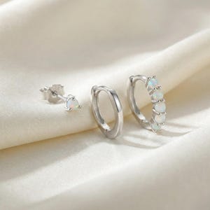 Può includere: Un set di tre orecchini d'argento. Uno è uno stud con una piccola pietra di opale. Gli altri due sono orecchini a cerchio, uno semplice e l'altro con cinque pietre di opale. Gli orecchini sono presentati su un tessuto bianco e setoso.