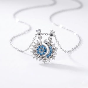 May include: A silver necklace featuring a sun and moon pendant. The sun has a blue center and radiating rays, while the moon is a crescent shape with blue accents. The pendant is adorned with small, clear crystals and hangs from a delicate chain.
