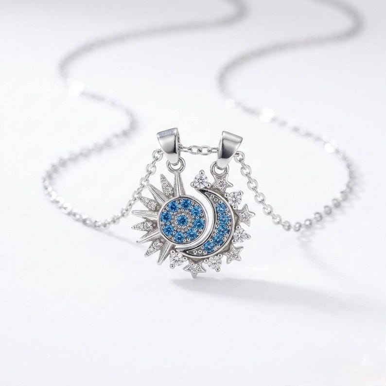 May include: A silver necklace featuring a sun and moon pendant. The sun has a blue center and radiating rays, while the moon is a crescent shape with blue accents. The pendant is adorned with small, clear crystals and hangs from a delicate chain.