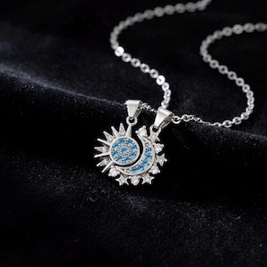 May include: A silver necklace featuring two pendants: a sun and a crescent moon. The sun pendant has a blue center with surrounding rays, while the moon pendant has a blue crescent with surrounding stars. Both pendants are adorned with small, clear stones.