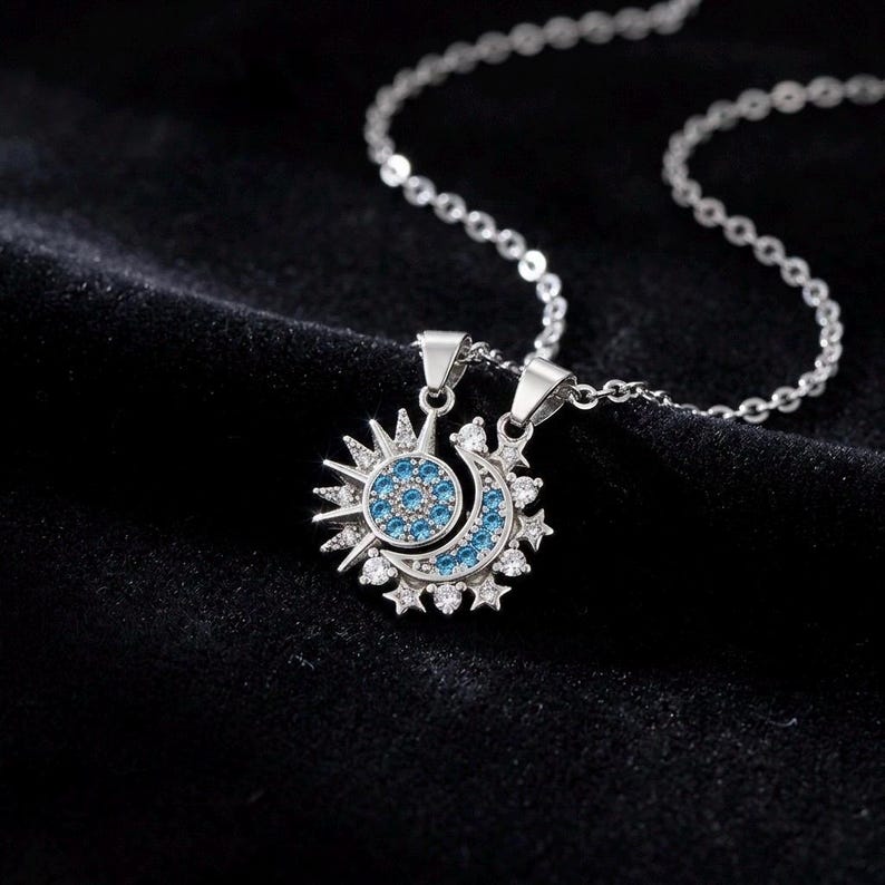 May include: A silver necklace featuring two pendants: a sun and a crescent moon. The sun pendant has a blue center with surrounding rays, while the moon pendant has a blue crescent with surrounding stars. Both pendants are adorned with small, clear stones.