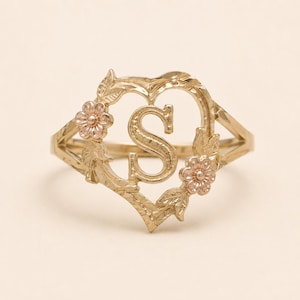 May include: A gold ring featuring a heart-shaped frame with a central letter S. The design includes textured leaves and two small flowers with rose-colored centers. The band is thin and tapers toward the back, showcasing a polished metallic finish.