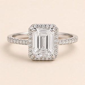 May include: A silver-toned ring featuring a large, rectangular emerald-cut center stone surrounded by a halo of small, round, clear stones. The band is slender and also encrusted with a row of small, round, clear stones along the top half.