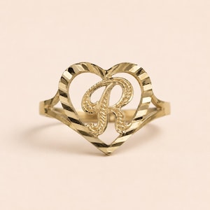 May include: A gold ring featuring a heart-shaped frame with a textured, diagonal striped finish. Centered within the heart is a large, ornate capital letter R with a decorative, swirling script design. The band is thin and tapers toward the back.
