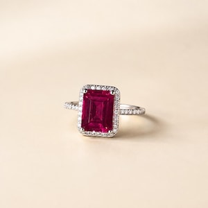 May include: A silver-toned ring featuring a large, rectangular, emerald-cut vibrant pinkish-red gemstone at its center. The central stone is surrounded by a halo of small, clear, round-cut stones, with additional small stones set along the thin band.