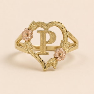 May include: A gold ring featuring a heart-shaped frame with a textured letter P in the center. The heart is decorated with two small rose gold flowers and leaves on the sides. The metal has a polished finish with intricate detailing on the surface.