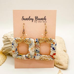 May include: Square earrings with a marbled design in white, blue, red, and gold. The earrings have gold-colored wire accents and hooks. The earrings are displayed on a pink card with the text "Sunday Brunch by the Sea".