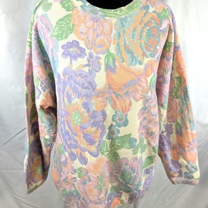 May include: A long-sleeved sweatshirt with a cream-colored background and a large floral pattern in shades of peach, lavender, light blue, and green. The crew neck and cuffs are trimmed in a light green color.