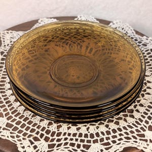 May include: A stack of three round amber-colored glass saucers featuring a textured, web-like pattern on the underside. The glass is translucent with a smooth, glossy finish on the top surface and a circular indentation in the center of each plate.