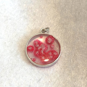 Puede incluir: Colgante redondo con bisel plateado. El colgante presenta resina transparente con purpurina roja y formas rojas en forma de burbujas. Tiene un pequeño lazo plateado en la parte superior para una cadena.