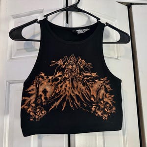 Puede incluir: Camiseta sin mangas negra con un diseño gráfico marrón que representa una figura con una esfera de reloj y calaveras. La camiseta sin mangas está colgada de una percha negra.