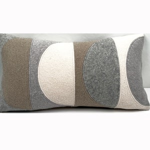 May include: Rectangular decorative pillow with a geometric design. The pillow features a patchwork of neutral colors, including light beige, gray, and brown. The design is composed of curved and rectangular shapes, creating a modern aesthetic. The pillow is made of a textured fabric.