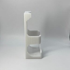 Puede incluir: Un organizador vertical blanco impreso en 3D con un dise&ntilde;o acanalado. Cuenta con dos estantes curvos y una secci&oacute;n superior, adecuado para guardar objetos peque&ntilde;os. El organizador est&aacute; hecho de pl&aacute;stico de color claro.