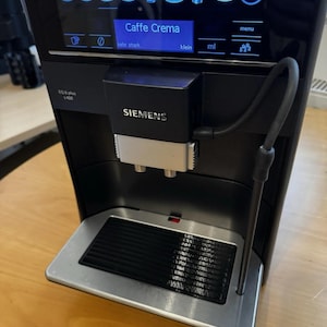 Puede incluir: Máquina de espresso Siemens EQ.6 plus s400 negra con una pantalla digital que muestra "Caffe Crema". La máquina tiene una bandeja de goteo plateada y una boquilla de vapor. La pantalla muestra varias opciones de bebidas de café.