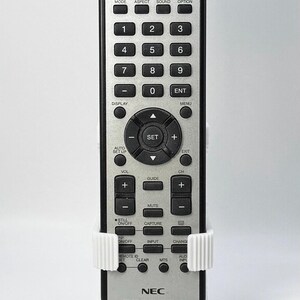 Puede incluir: Mando a distancia NEC plateado con numerosos botones, incluyendo controles de encendido, volumen y canal. El mando se sujeta en un soporte blanco impreso en 3D. El n&uacute;mero de modelo RU-M117 es visible.