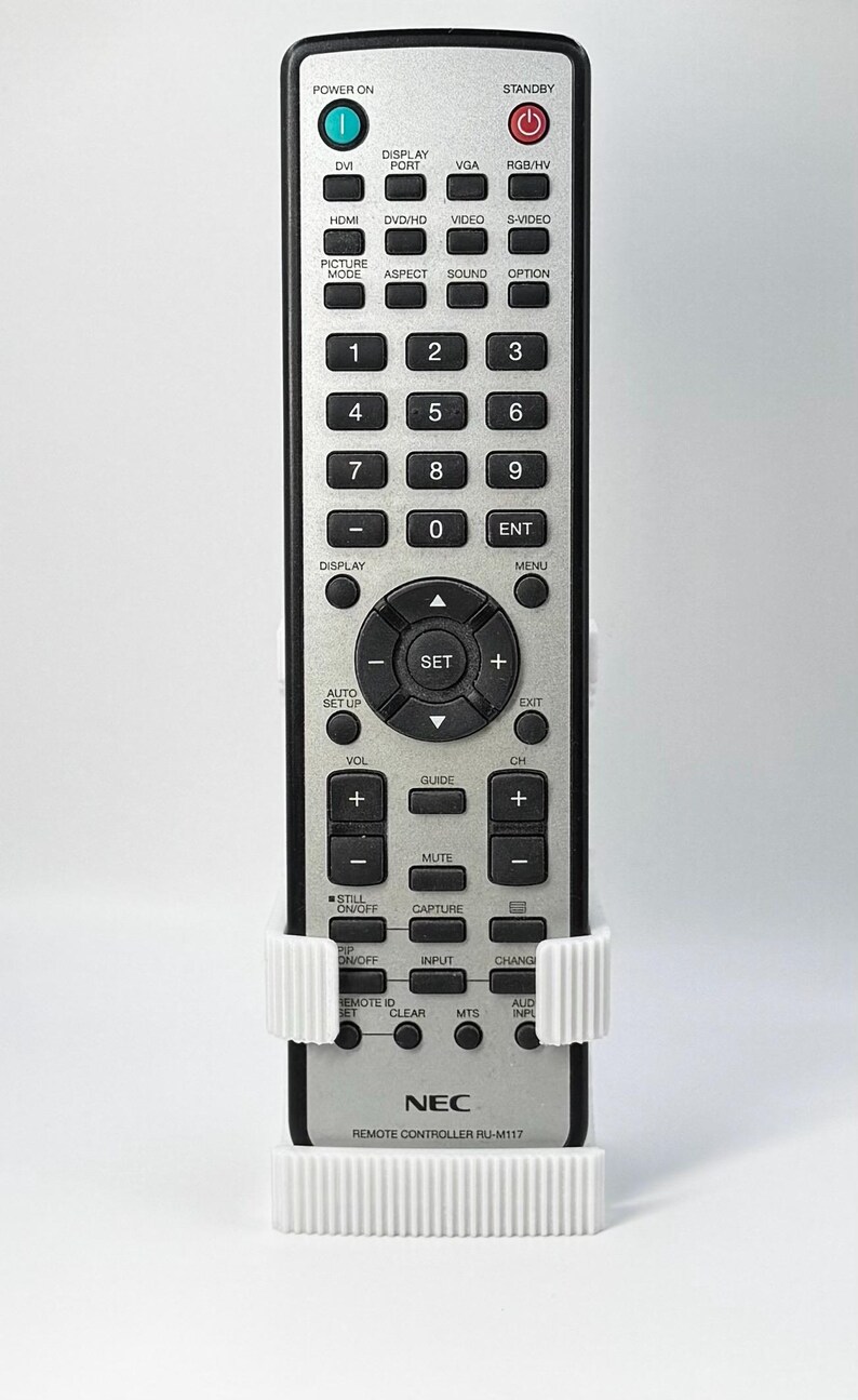 Puede incluir: Mando a distancia NEC plateado con numerosos botones, incluyendo controles de encendido, volumen y canal. El mando se sujeta en un soporte blanco impreso en 3D. El n&uacute;mero de modelo RU-M117 es visible.