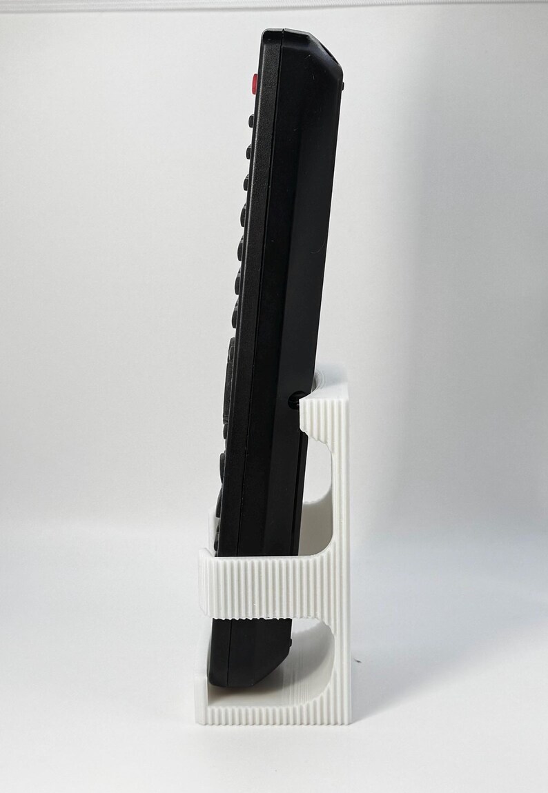 Puede incluir: Un mando a distancia negro con m&uacute;ltiples botones y un bot&oacute;n rojo de encendido se coloca en un soporte blanco, acanalado, impreso en 3D. El soporte tiene dos aberturas y una base rectangular.