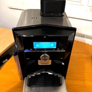 Puede incluir: Una cafetera automática Siemens EQ.9 negra y plateada con una pantalla digital que muestra "Cappuccino". La máquina tiene una parte superior plateada, un panel frontal negro y una bandeja de goteo plateada. Un recipiente negro se encuentra encima.