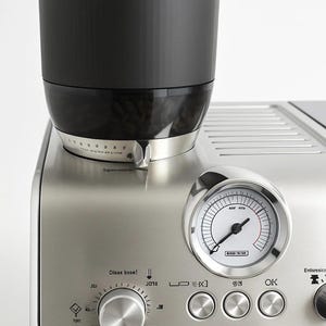 Puede incluir: Máquina de espresso de acero inoxidable con un molinillo de granos negro en la parte superior. La máquina tiene un manómetro y perillas de control. La marca "De'Longhi" es visible.