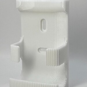 Puede incluir: Un soporte de pared blanco, impreso en 3D, con un cuerpo rectangular. Cuenta con una abertura ovalada central y dos brazos curvos y acanalados que se extienden desde los lados. La base tambi&eacute;n tiene un dise&ntilde;o acanalado. El soporte est&aacute; dise&ntilde;ado para sujetar un objeto.