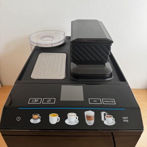Puede incluir: Máquina de espresso Siemens negra con molinillo incorporado y varias opciones de bebidas. La máquina tiene una pantalla digital y botones para diferentes tipos de café, incluyendo espresso, capuchino y latte. Sobre una superficie de madera.