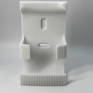 Puede incluir: Un objeto blanco impreso en 3D con forma rectangular. Cuenta con dos ranuras empotradas y dos soportes laterales con estr&iacute;as verticales. El borde inferior tiene una serie de estr&iacute;as paralelas. El objeto est&aacute; dise&ntilde;ado para sujetar algo.