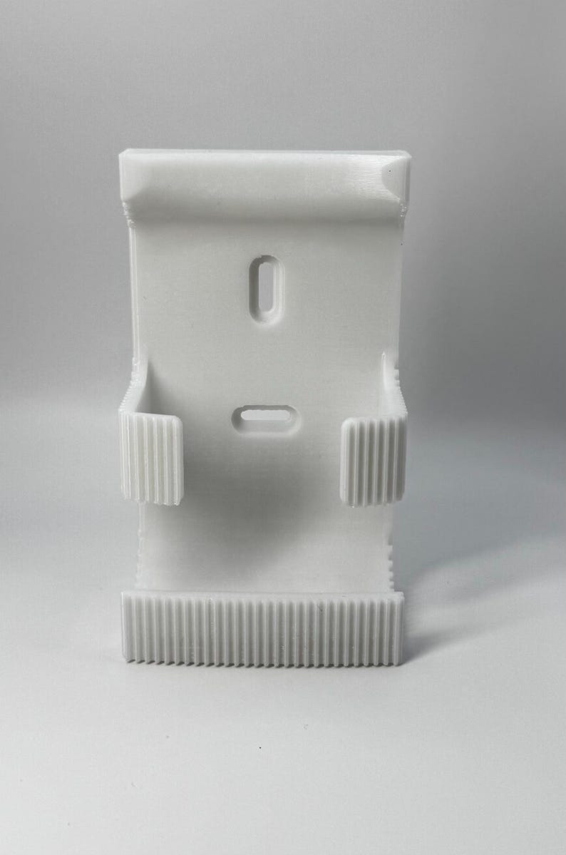 Puede incluir: Un objeto blanco impreso en 3D con forma rectangular. Cuenta con dos ranuras empotradas y dos soportes laterales con estr&iacute;as verticales. El borde inferior tiene una serie de estr&iacute;as paralelas. El objeto est&aacute; dise&ntilde;ado para sujetar algo.